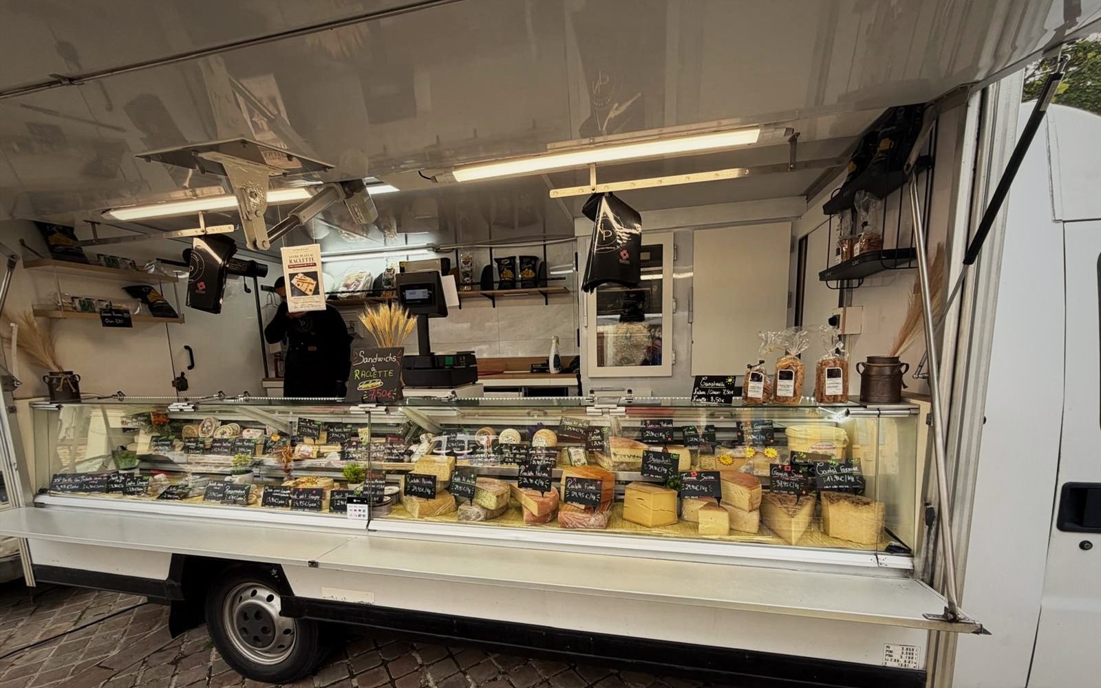 Le camion-étal NP Fromages ouvert au marché, sa vitrine garnie de fromages et d’ardoises de prix